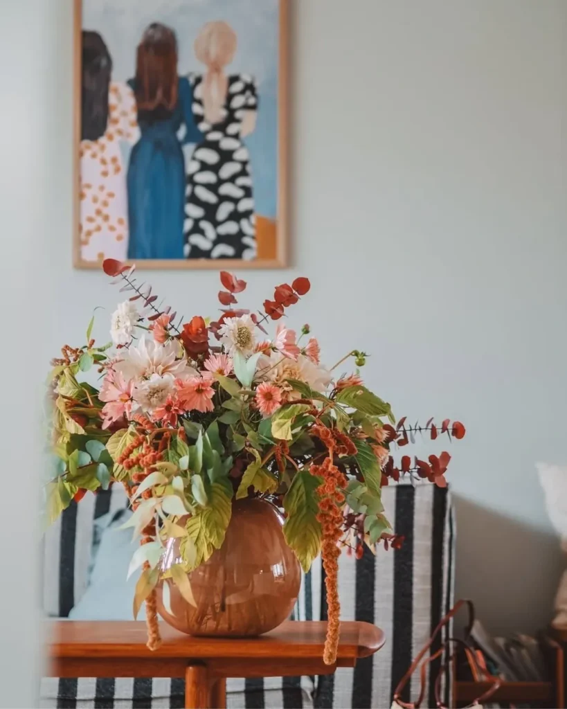 Imagem com vaso de flores e ao fundo um quadro com três mulheres