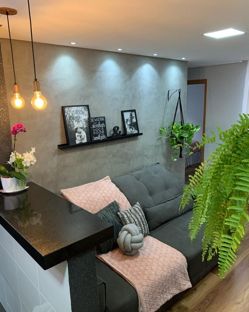 Sala de estar com sofá cinza e manta rosa, prateleira preta com quadrinhos