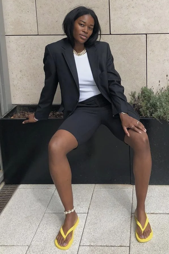 Mulher de pele preta, com cabelo escuro, look de alfaiataria e chinelo de dedo amarelo