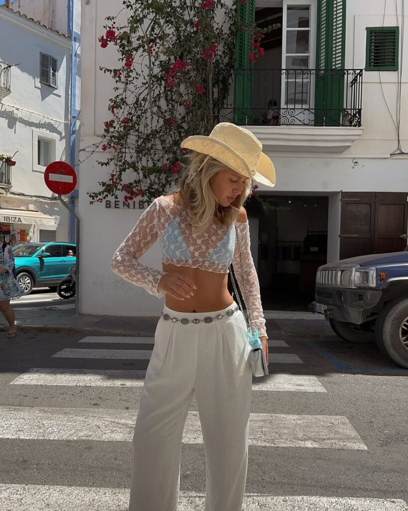 Mulher de pele clara, com calça branca, blusa de renda e chapéu country