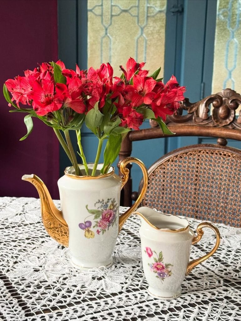 Mesa com toalha de crochê branca e bules com estampas florais e detalhes dourados