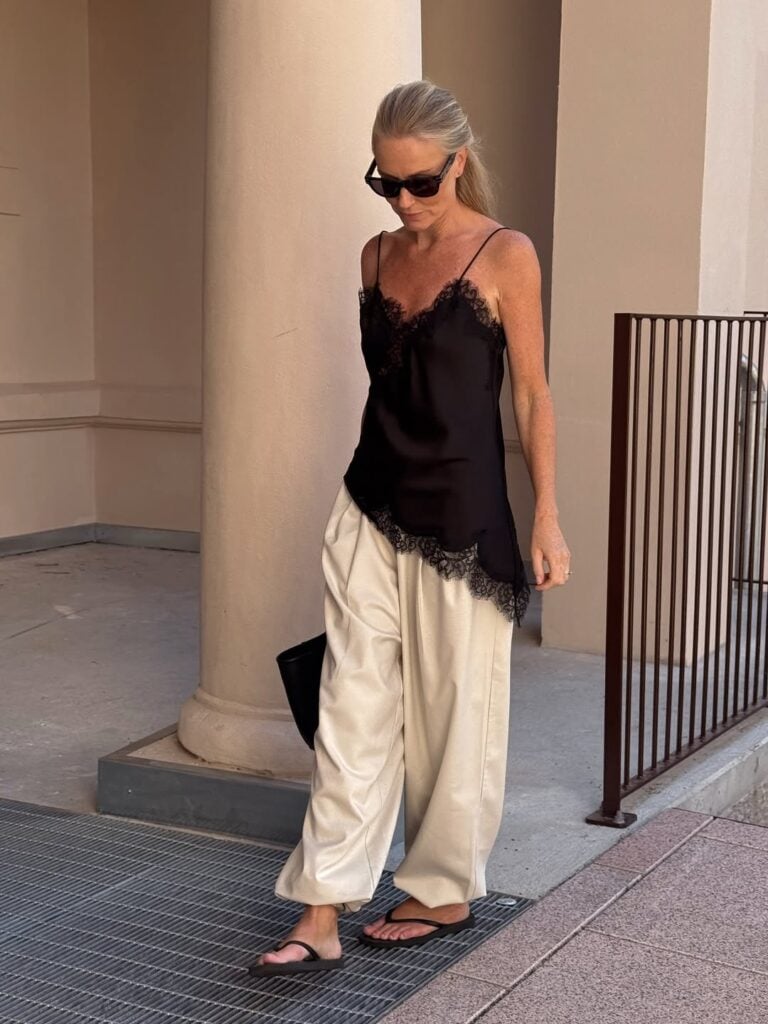 Mulher de pele clara, com blusa assimétrica de renda, calça clara, bolsa de mão e chinelo de dedo