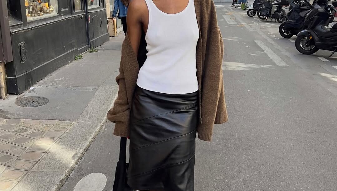 Mulher jovem e magra com pele escura. Ela usa regata branca e saia de couro preta.