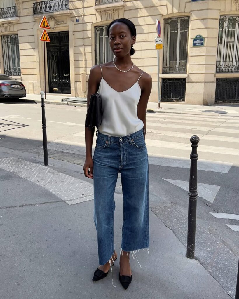 Mulher jovem e magra com look de escritório com blusa de cetim, calça jeans e salto alto
