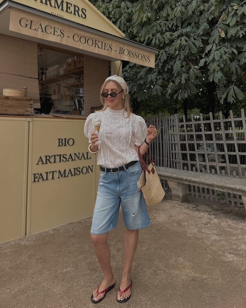Mulher de pele clara, com blusa branca rendada, jorts, bandana no cabelo e chinelo de dedo
