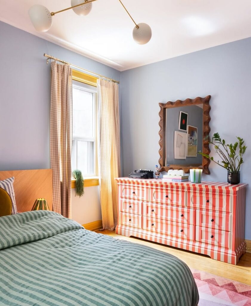 Quarto com paredes azul-claro, cômoda listrada em rosa e branco e espelho com moldura de madeira