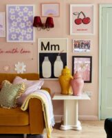 Parede rosa-claro com quadros, porta verde-claro, mesa lateral branca com vasos rosa e amarelo
