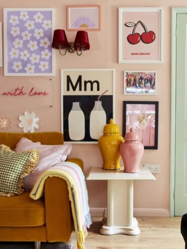 Parede rosa-claro com quadros, porta verde-claro, mesa lateral branca com vasos rosa e amarelo