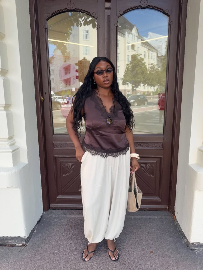 Mulher de pele preta, com calça branca aladin e regata marrom de renda