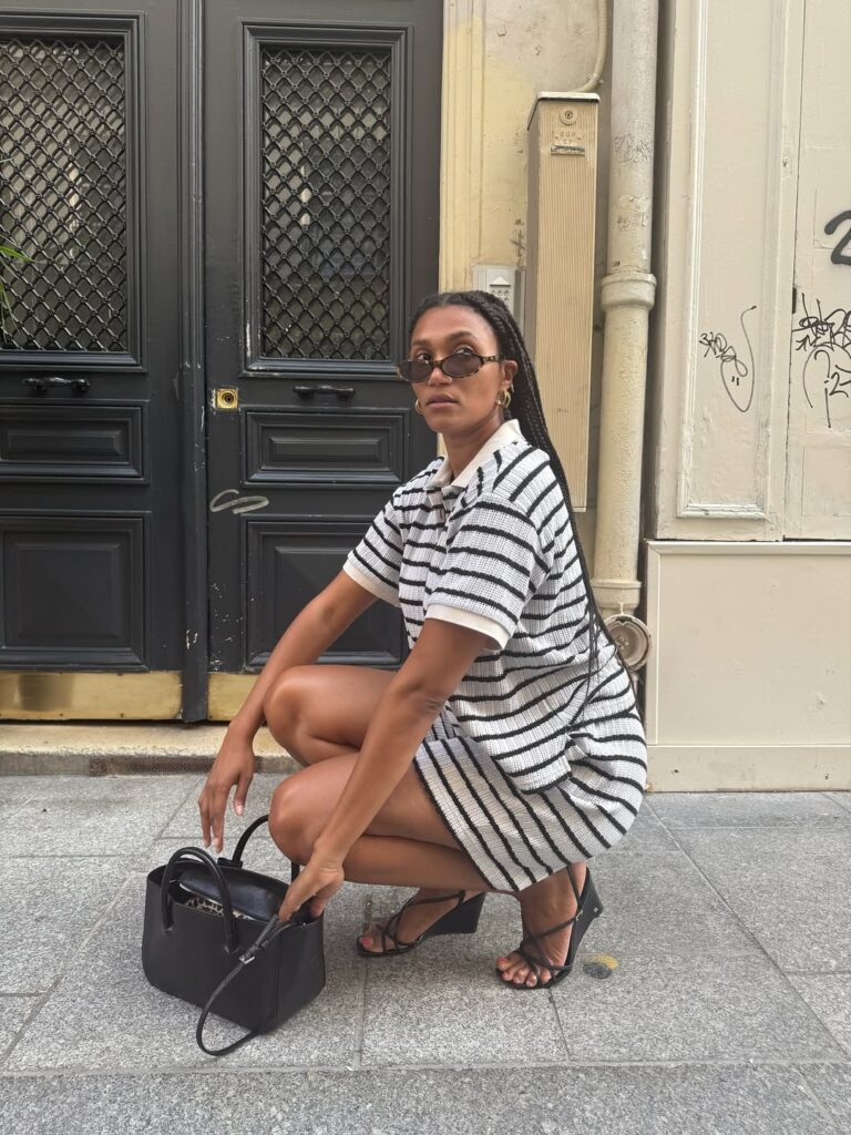 Mulher de pele preta, com vestido listrado, bolsa preta e sandália anabela escura
