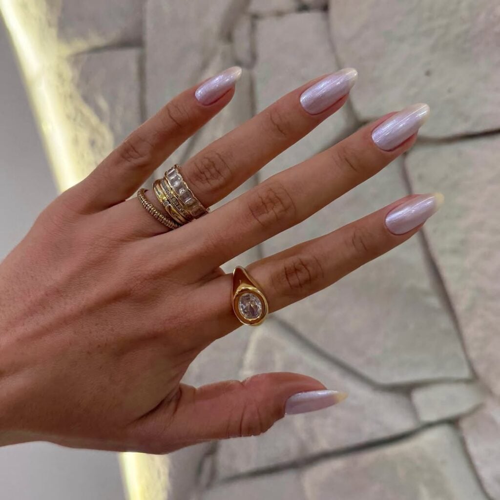 Mão de pele clara, com anéis em dourado e unha amendoada branca e perolada