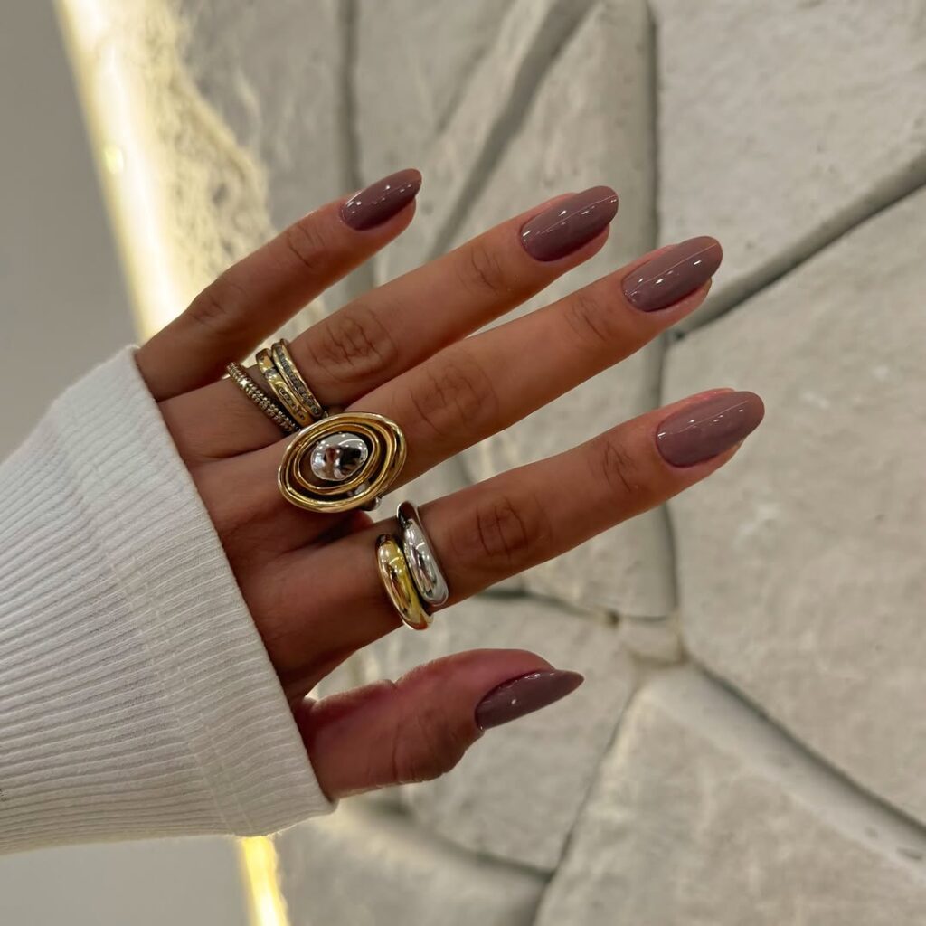 Mão de pele clara, com anéis em prata e dourado e unha amendoada nude acinzentado