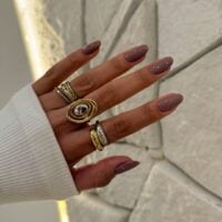 Mão de pele clara, com anéis em prata e dourado e unha amendoada nude acinzentado