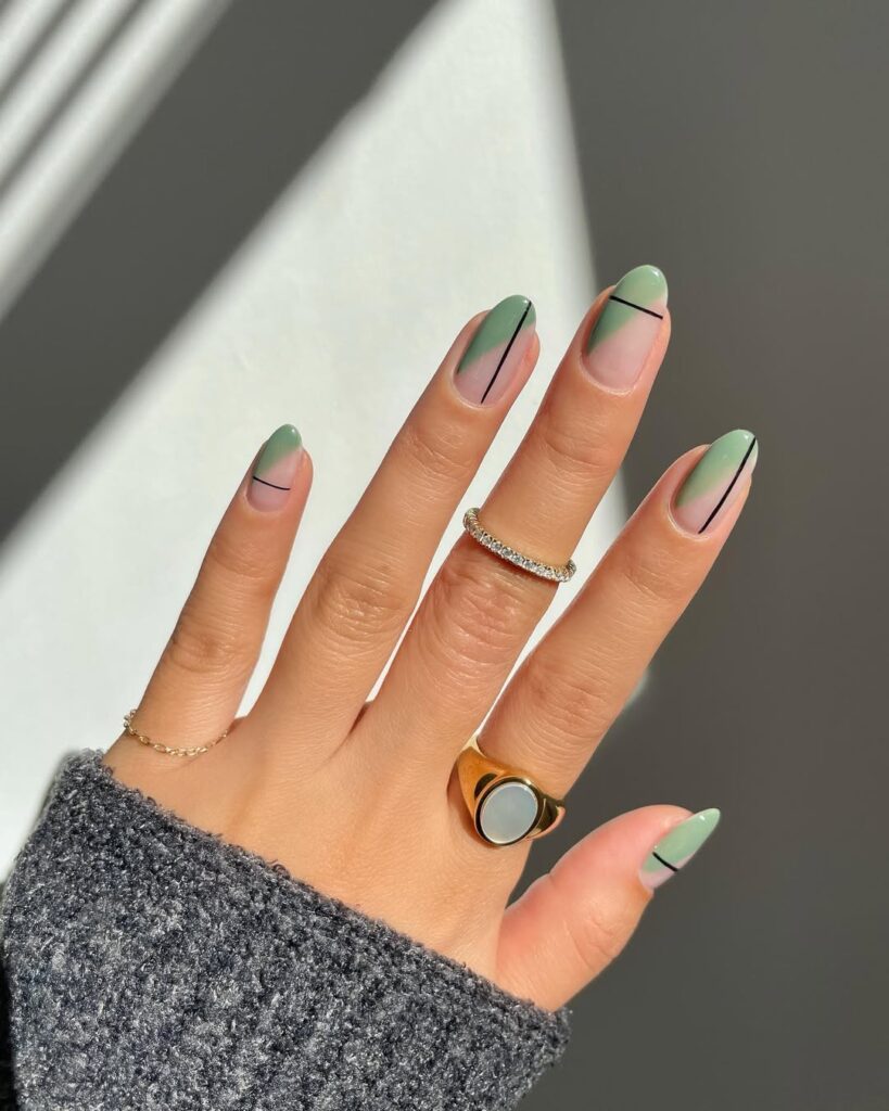 Mão de pele clara, com anéis em dourado e unha amendoada verde com linhas em preto 