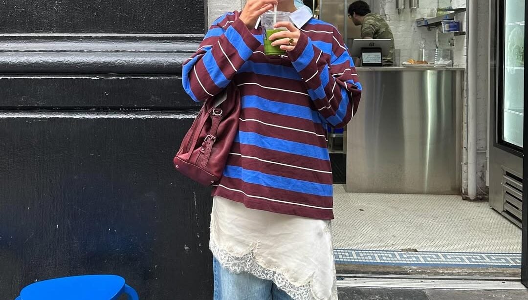 mulher jovem e magra com blusa de listras, vestido acetinado por cima da calça jeans e sapatilha