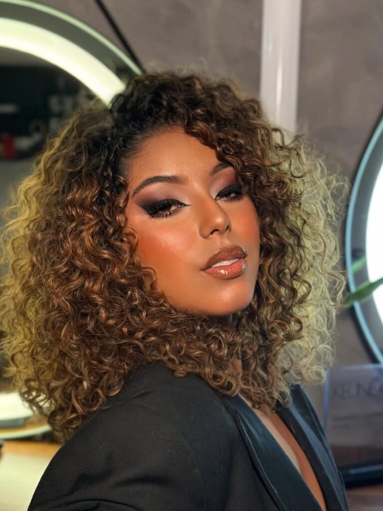 Mulher de pele preta, com blazer e maquiagem marcante. Ela tem o cabelo cacheado iluminado