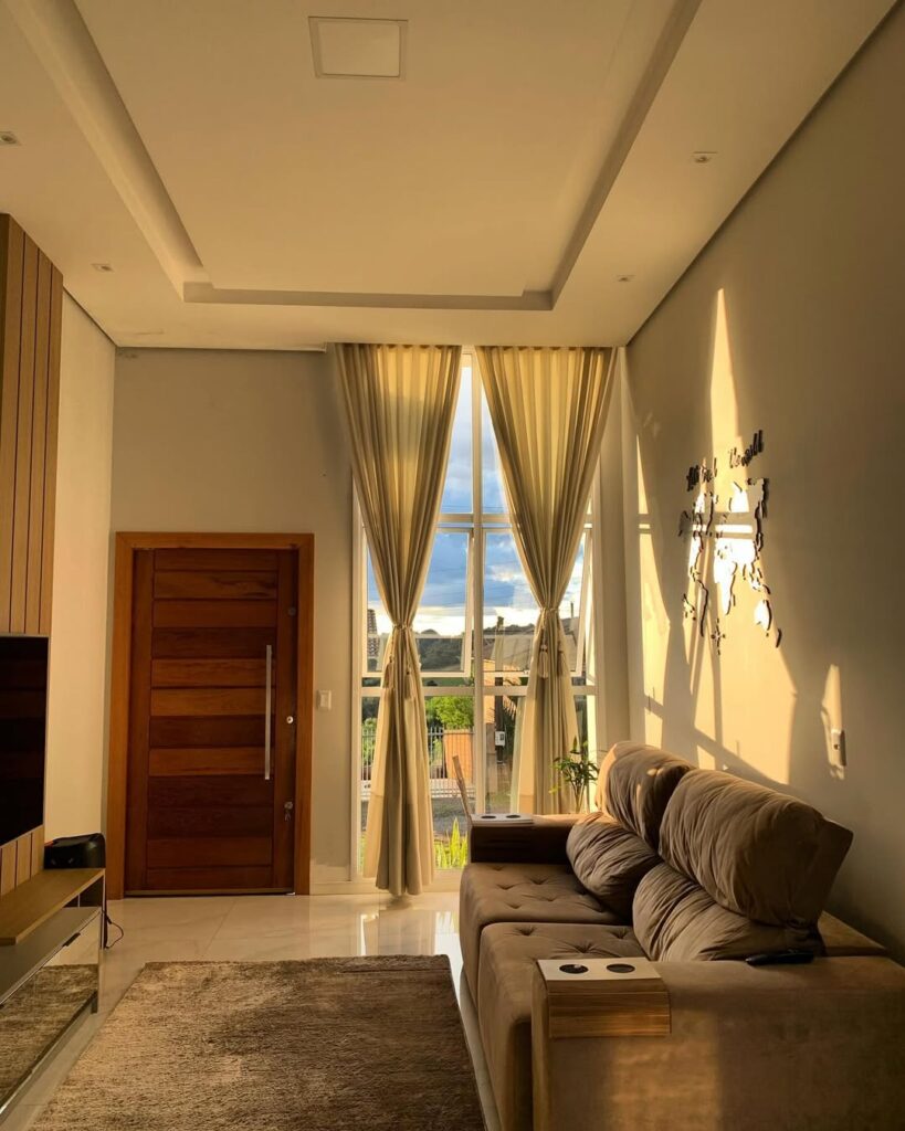 Sala de estar com porta de madeira, teto de gesso e grande janela com cortina