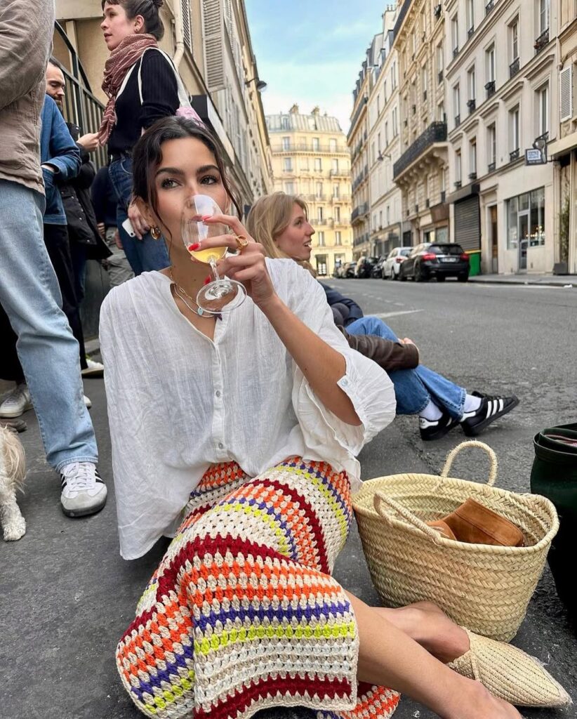 Mulher de pele clara, sentada no chão tomando vinho, com camisa branca e saia de croche