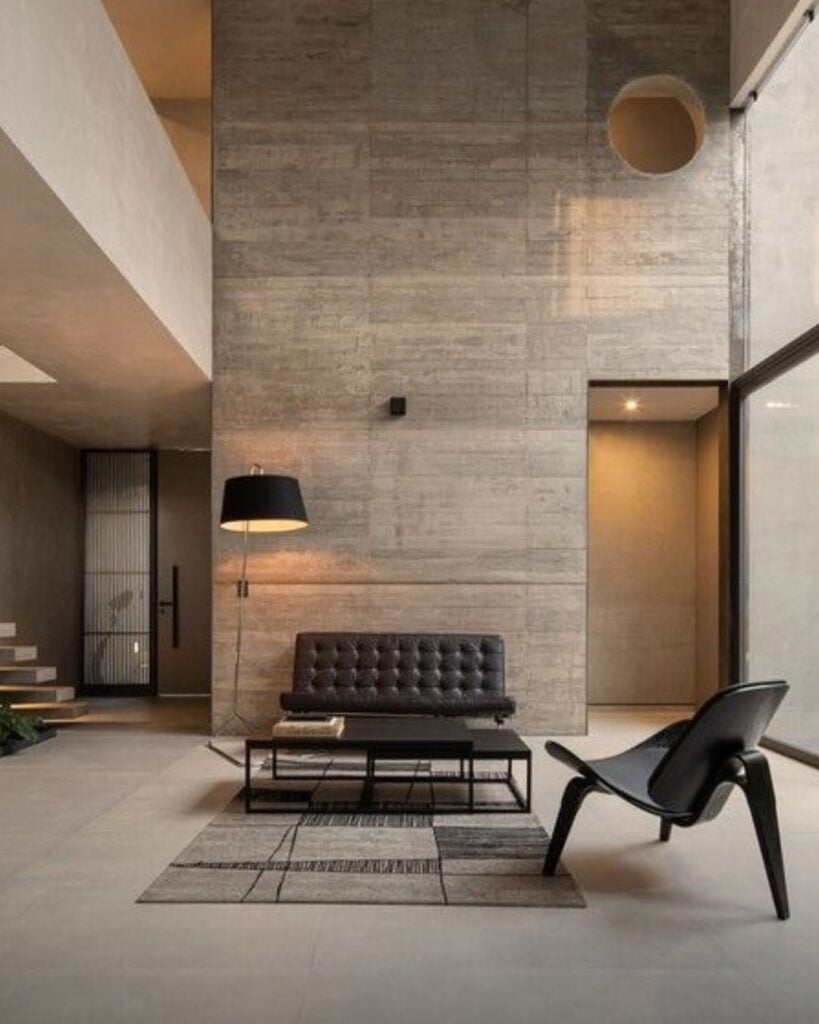 Sala de estar com sofá, mesa de centro e poltrona pretos, luminária de piso e parede revestida com concreto