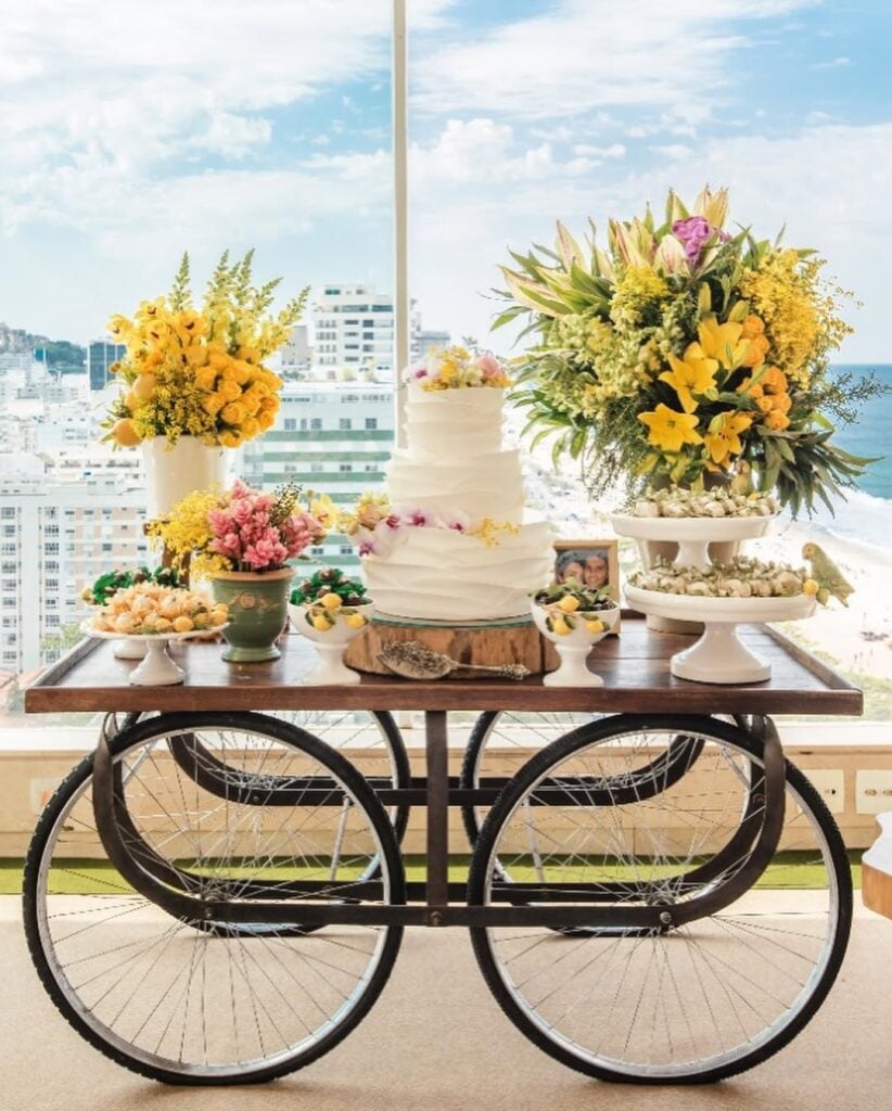 Mesa de madeira com rodas de bicicleta, bolo branco de três andares e arranjos com flores amarelas
