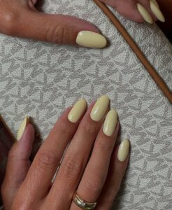 Mãos de pele clara, com anéis em dourado e unha amendoada amarelo-manteiga