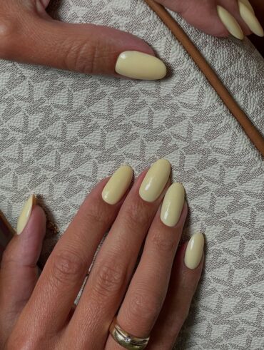 Mãos de pele clara, com anéis em dourado e unha amendoada amarelo-manteiga