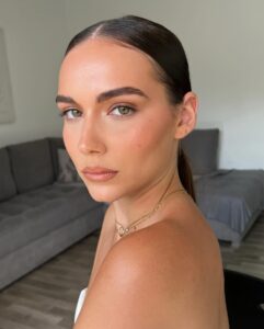 Mulher de pele clara, com acessórios em dourado e cabelo preso. Ela está com uma maquiagem clean