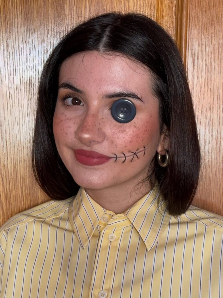 Mulher de pele clara, com cabelo escuro curto, camisa amarela e maquiagem de Coraline
