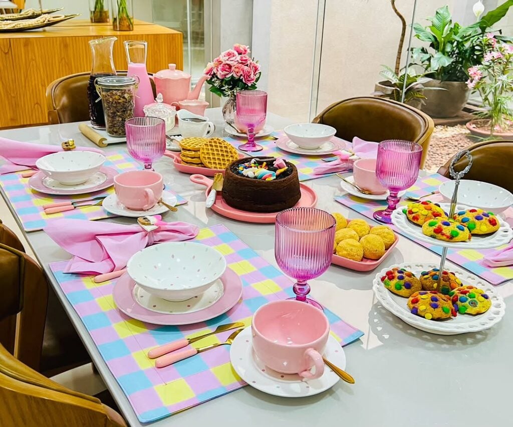 Mesa posta com jogo americano em tons pastel, copos rosa e bolo de chocolate