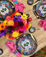 Mesa posta de dia de los muertos com sousplats de caveiras, talheres dourados e flores coloridas