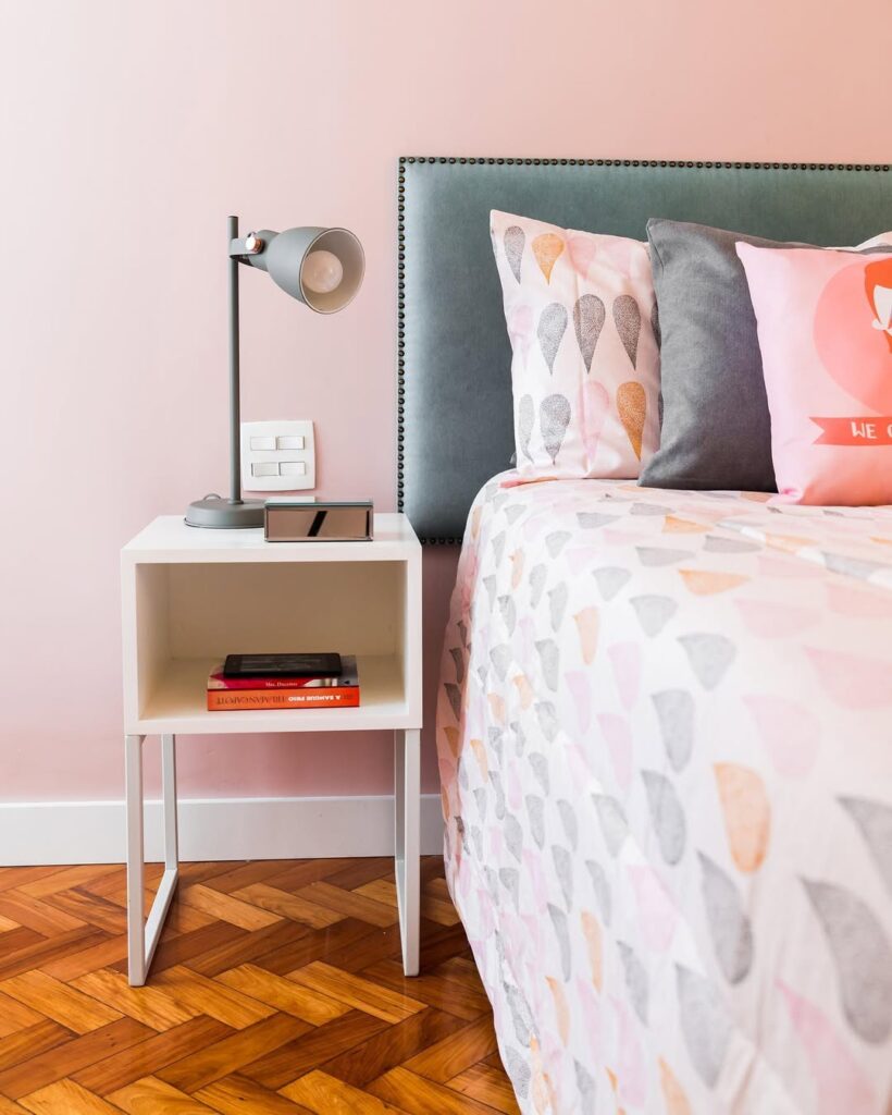 Cama com cabeceira cinza e mesa de cabeceira branca com livros e luminária