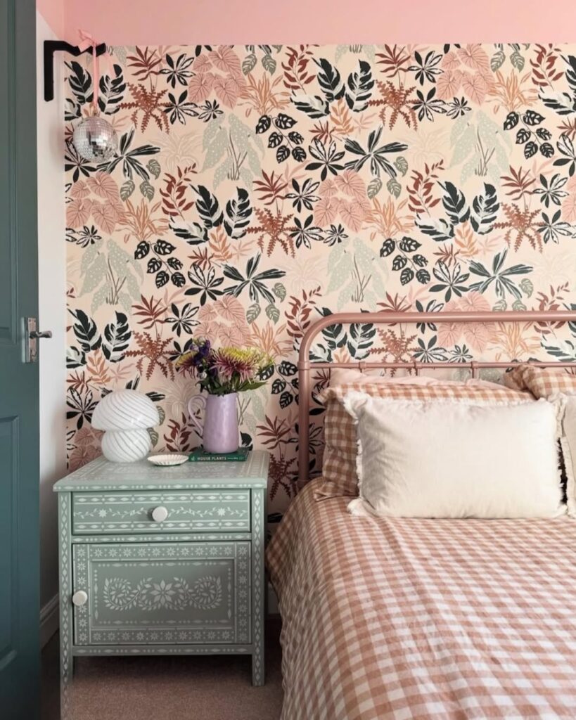 Quarto com cama de ferro em tom de rosa, mesa de cabeceira verde-claro com detalhes brancos e papel de parede floral