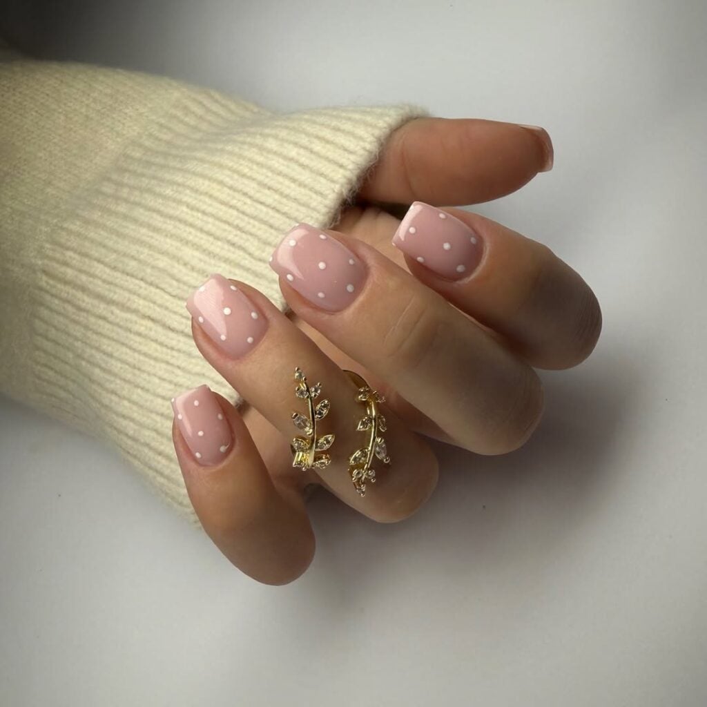 Mão de pele clara, com anel em dourado e unha quadrada nude com poá branco