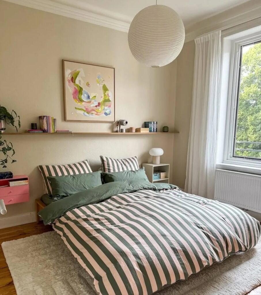 Quarto de casal com roupa de cama listrada em branco e verde, prateleira com objetos decorativos e luminária japonesa