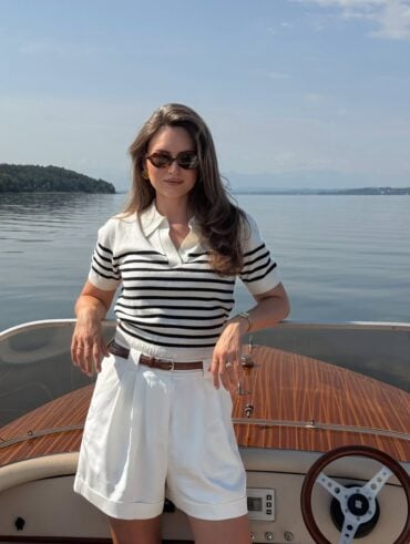 Mulher de pele clara, com cabelo castanho, blusa de listras, short branco e óculos de sol