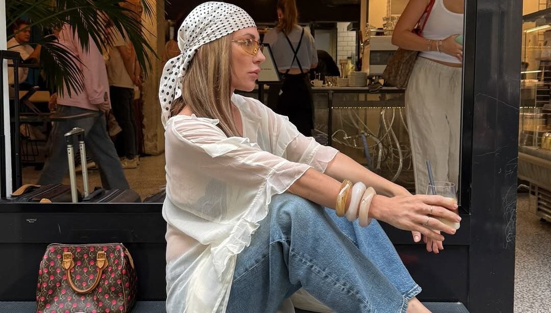 Mulher jovem sentada em banco. Ela usa blusa alongada, calça jeans, lenço na cabeça e calçados vermelhos