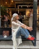 Mulher jovem sentada em banco. Ela usa blusa alongada, calça jeans, lenço na cabeça e calçados vermelhos