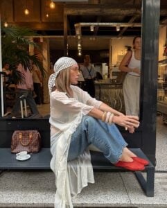 Mulher jovem sentada em banco. Ela usa blusa alongada, calça jeans, lenço na cabeça e calçados vermelhos