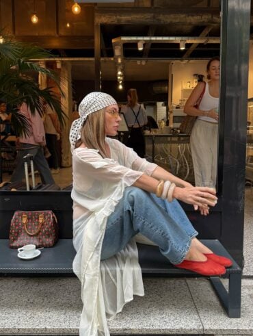 Mulher jovem sentada em banco. Ela usa blusa alongada, calça jeans, lenço na cabeça e calçados vermelhos