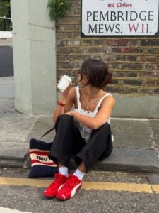 Mulher jovem em uma calçada. Ela usa calça preta, blusa branca e speedcat vermelho