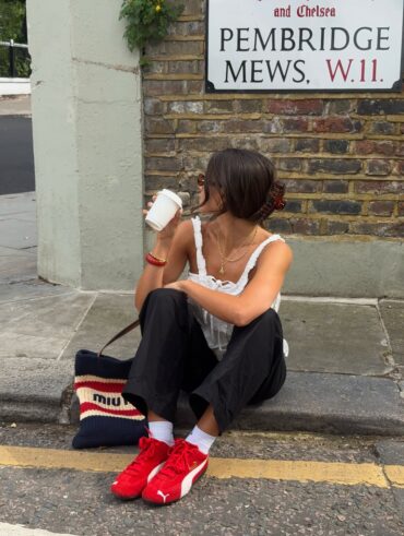 Mulher jovem em uma calçada. Ela usa calça preta, blusa branca e speedcat vermelho