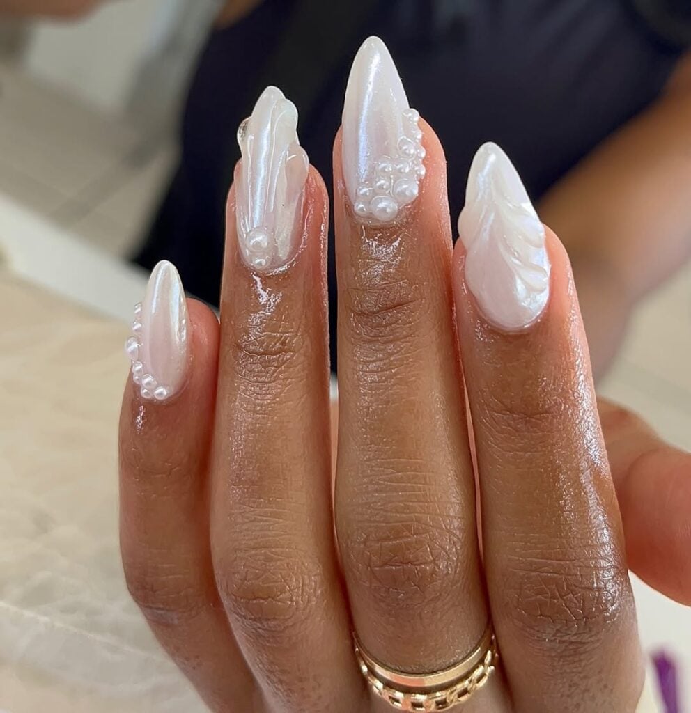 Mão de pele preta, com anéis em dourado e unha amendoada branca perolada com decorações com pérolas e gel de unha