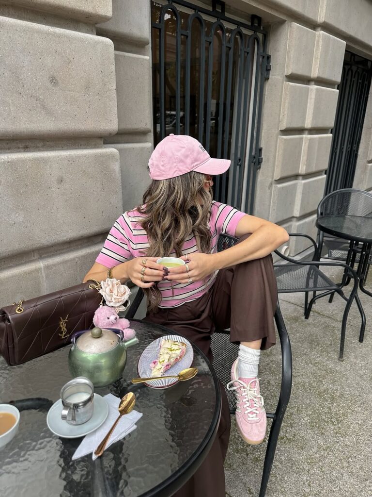 Mulher jovem e magra com pele clara sentada em uma cadeira na calçada. Ela usa blusa listrada rosa, boné e tênis rosa e calça marrom