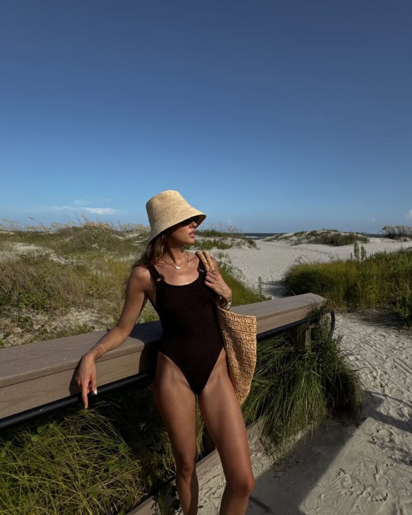 Mulher jovem e magra na praia. Ela usa maiô e chapéu de palha.