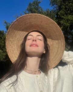 Anne Hathaway com blusa branca, cabelo castanho e chapéu de palha