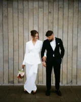 Casal com roupa de casamento. O homem usa blazer e calça social e camisa. A mulher usa conjunto de terno branco.