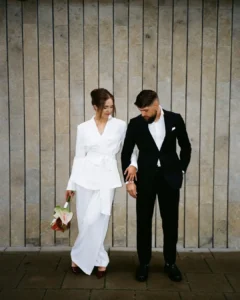 Casal com roupa de casamento. O homem usa blazer e calça social e camisa. A mulher usa conjunto de terno branco.