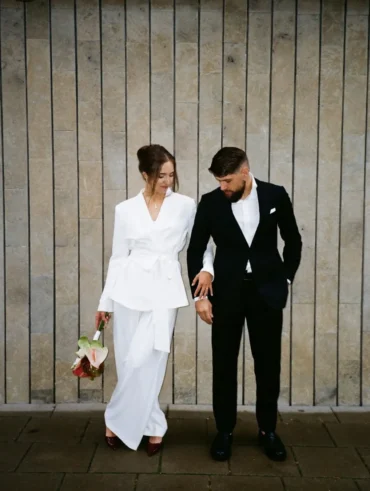 Casal com roupa de casamento. O homem usa blazer e calça social e camisa. A mulher usa conjunto de terno branco.