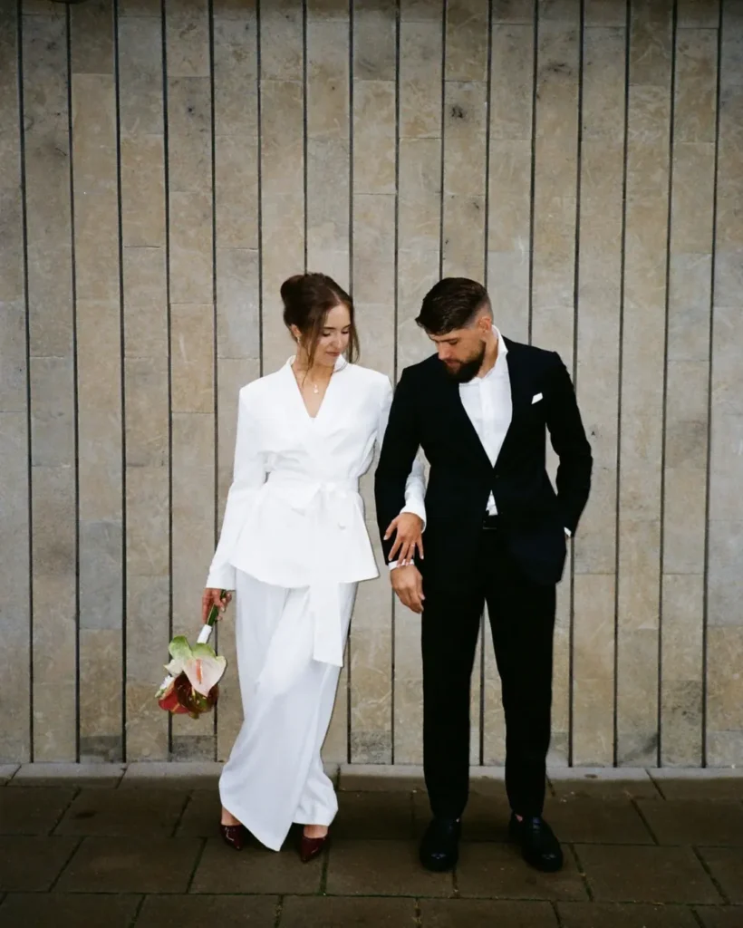 Casal com roupa de casamento. O homem usa blazer e calça social e camisa. A mulher usa conjunto de terno branco.