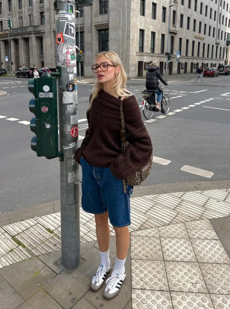 Mulher jovem usando suéter, bermuda jeans, tênis Adidas Samba com meia branca e bolsa de oncinha
